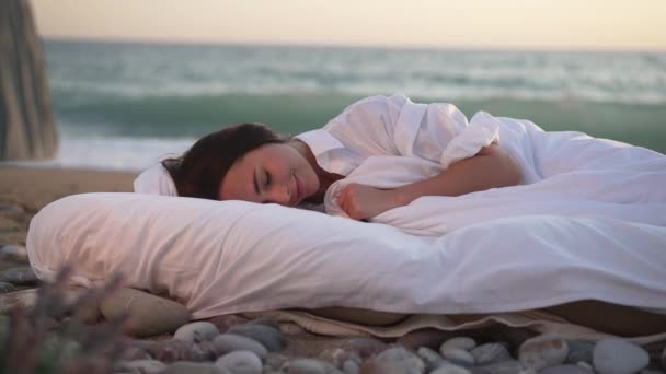 Vrolijke jonge lachende vrouw slapen in wit zacht bed met turquoise zee golven spatten met schuim op het zandstrand op de achtergrond. Portret van een zorgeloze blanke dame met gesloten ogen die buiten ligt. — Stockvideo