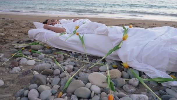 Mulher bonita relaxada sorrindo alongando-se na cama confortável almofadada com flores amarelas espalhadas e espumosas ondas turquesa rolando em câmera lenta no fundo. Retrato de senhora caucasiana feliz. — Vídeo de Stock