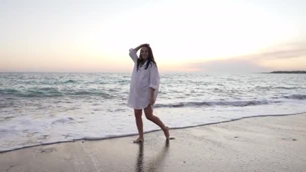 Ampia ripresa di sorridere a piedi nudi giovane donna magra guardando la fotocamera camminare in onde oceaniche schiumose al rallentatore. Ritratto di turista caucasico felice in posa al tramonto sulla costa del mare all'aperto. — Video Stock