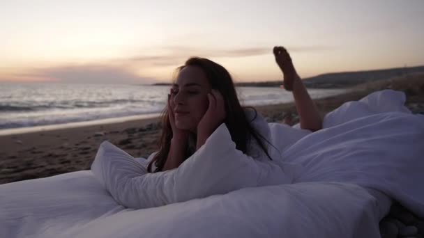 Молодая стройная улыбающаяся женщина, лежащая на мягкой кровати в сумерках с живописным средиземноморским пейзажем на заднем плане. Портрет счастливой брюнетки, отдыхающей на свежем воздухе в сумерках. — стоковое видео