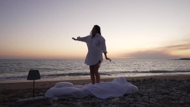 Siluet tembakan lebar dari wanita ramping Kaukasia yang bersemangat melompat dan berputar dalam gerakan lambat di pantai laut Mediterania yang indah saat senja. Wanita milenial gembira bersenang-senang di senja hari di pantai laut. — Stok Video