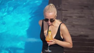Sarışın, zayıf, yetişkin bir kadın turistin havuz kenarında gün ışığında ağır çekimde portakal kokteyli içişini yüksek açıdan görmek. Yaz tatilinde dinlenen kendine güvenen muhteşem bir kadının portresi..