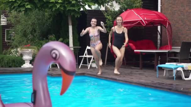 Amplio tiro dos mujeres jóvenes y alegres caucásicas corriendo saltando en la piscina con agua azul azur salpicando en cámara lenta. Retrato de turistas positivos amigos divirtiéndose en el resort al aire libre. — Vídeos de Stock