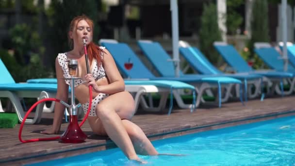 Amplio retrato de foto de una hermosa mujer caucásica de talla grande sentada con los pies en el agua de la piscina fumando narguile en cámara lenta. Relajado despreocupado hermoso turista disfrutando de la relajación al aire libre bajo el sol. — Vídeo de stock