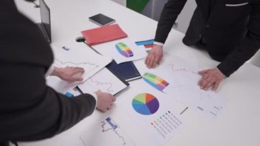 Ofisinde grafikler olan yüksek açı masası ve iki konsantre erkek meslektaşı evrak işlerini inceliyor. Kapalı alanda çalışan kendine güvenen beyaz adamlar yavaş çekimde iş stratejisi planlıyorlar.