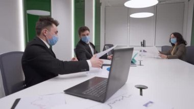 Coronavirus 'taki iş ekibi maske takıp konferans masasına oturmuş evrak işlerini inceliyor. Covid-19 salgınında ofiste çalışan kendinden emin profesyonel erkek ve kadın..