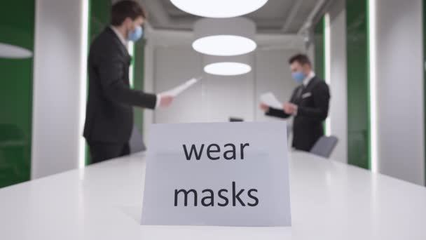 Wear masks message in business office with blurred Caucasian men in coronavirus face masks passing documents at background. Social distancing and Covid-19 prevention on pandemic outbreak. — Stock Video
