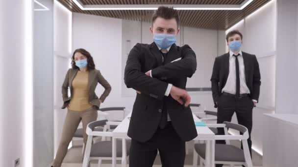 Portrait d'un homme caucasien confiant croisant les mains en regardant la caméra avec des collègues flous dans le masque facial coronavirus en arrière-plan. Des hommes d'affaires sérieux posant au bureau sur Covid-19 lockdown. — Video