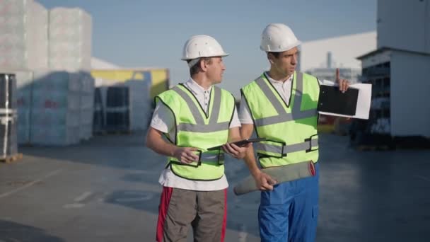 Portret van serieuze zelfverzekerde Midden-Oosten en Kaukasische ingenieurs die in het zonlicht buiten in het magazijn praten over verzending. Professionele mannen in harde hoeden plannen lading levering. — Stockvideo