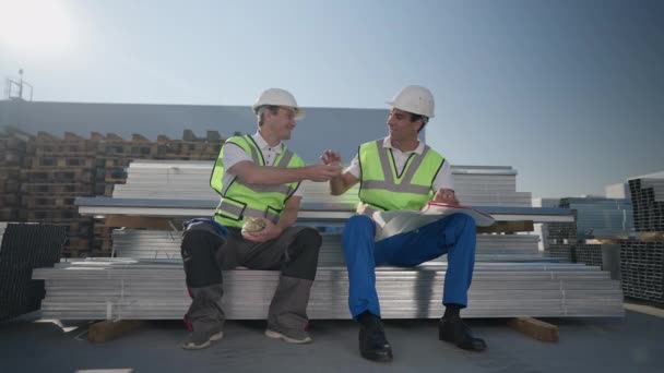 Wide shot portrait of positive Caucasian and Middle Eastern workers sharing lunch sitting outdoors at warehouse. Smiling handsome men talking shaking hands eating sandwich in slow motion. — Stock Video