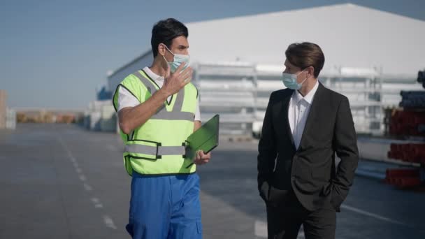 Serious Middle Eastern warehouse worker in uniform and Caucasian supervisor in suit walking talking outdoors on coronavirus pandemic. Portrait of professional men in Covid face masks discussing plan. — Stock Video