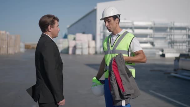 Vista laterale di arrabbiato travolto uomo del Medio Oriente passando uniforme al manager caucasico in giacca e cravatta parlando al rallentatore. Ritratto di impiegato di magazzino oberato di lavoro che litiga con il datore di lavoro all'aperto. — Video Stock