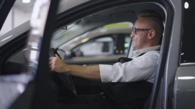 Gözlüklü, kulağı delinmiş, kendine güvenen, iç mekanları inceleyen başarılı beyaz adamın yan görüntüsü. Otomobili tercih eden varlıklı bir müşteri. Yavaş çekim.