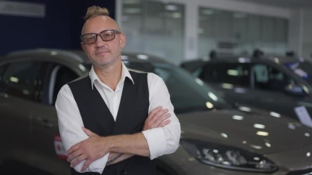 Portrait of proud wealthy Caucasian man in eyeglasses standing in car dealership looking at camera. Confident handsome rich buyer posing in auto showroom indoors. Slow motion. — Stock Video