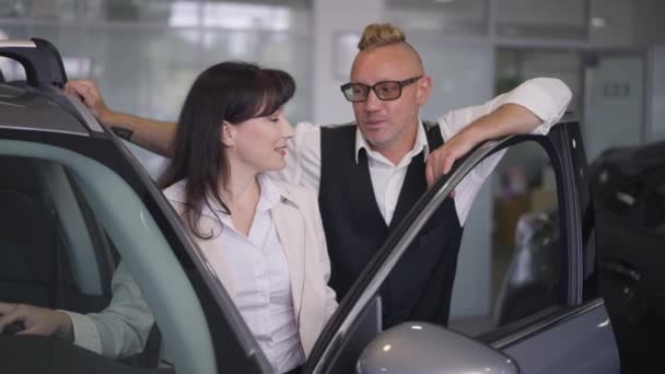 Pareja caucásica adulta cariñosa que está parada en concesionario del coche discutiendo opción del vehículo. Hombre guapo y hermosa mujer hablando sonriente elegir automóvil en sala de exposición en cámara lenta. — Vídeos de Stock