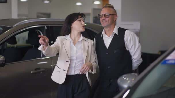 Beautiful Caucasian woman walking with handsome loving man in car dealership talking. Live camera follows smiling couple choosing new vehicle indoors in showroom. Slow motion. — Stock Video