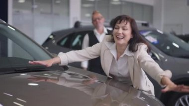 Heyecanlı güzel bir kadının portresi yepyeni lüks bir arabaya dokunuyor ve arka planda sevgili bir adam gülümsüyor. Mutlu eş, kapalı mekanda satılmasına şaşırdı. Yavaş çekim.