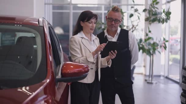 Araba satıcısı ve alıcının portresi galerideki yeni lüks kırmızı aracın önünde ağır çekimde konuşuyor. Profesyonel Kafkas uzmanı müşterinin kapalı mekanda otomobil seçmesine yardım ediyor.. — Stok video