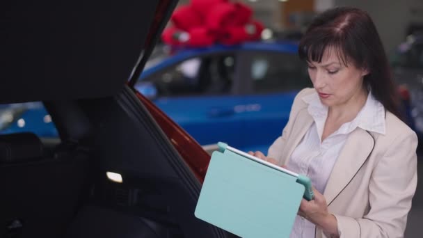 Профессиональная серьезная женщина-дилер автомобилей изучает новый роскошный автомобиль в дилерском центре с помощью планшета. Портрет великолепной уверенной в себе женщины, работающей в автосалоне. Медленное движение. — стоковое видео