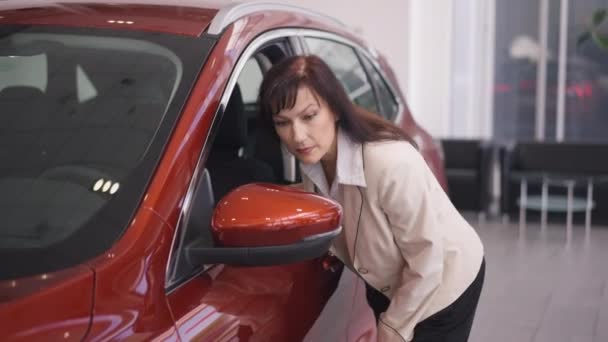 Mulher morena bonita olhando para o espelho do carro escolhendo novo veículo na concessionária. Retrato de confiante sério comprador caucasiano feminino examinando automóvel vermelho em câmera lenta no showroom. — Vídeo de Stock