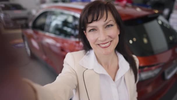 Selfie POV if cheerful beautiful female blogger streaming online from car dealership choosing new vehicle. Portrait of positive confident Caucasian adult woman blogging selecting automobile. — Stock Video