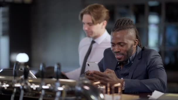 Rack εστίαση από χαρούμενα Αφρικής Αμερικανός άνθρωπος χαμογελώντας surfing Internet στο smartphone σε προβληματική Καυκάσιος τύπος που εισέρχονται μπαρ παραγγελία ποτών. Θετικός διευθυντής και αγχωμένος άνθρωπος στο εστιατόριο μετά την εργασία. — Αρχείο Βίντεο
