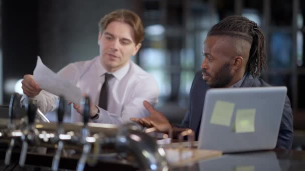 Un uomo afroamericano sicuro seduto al bar con un collega caucasico nervoso che discute l'idea del progetto. Ritratto di due manager che discutono di strategia aziendale al chiuso. Stile di vita e lavoro di squadra. — Video Stock