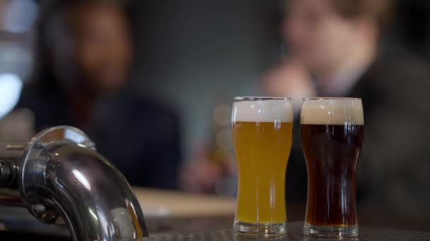 Primer plano lager pálido y oscuro en vasos en el mostrador de la barra con los hombres caucásicos y afroamericanos borrosas tostando botellas tintineo beber en el fondo. Concepto de servicio e industria del alcohol. — Vídeo de stock