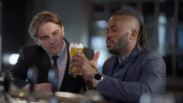 Afrikalı Amerikalı bira uzmanı, beyaz bir arkadaşla konuşuyor ve bardağı işaret eden soluk bira yüzlerini anlatıyor. İşten sonra barda dinlenen takım elbiseli konsantre adamların portresi.. — Stok video