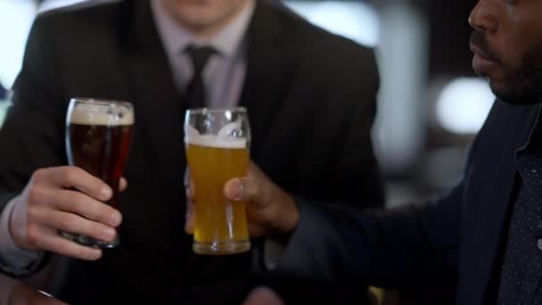 Primer plano afroamericanos y caucásicos hombres tintineo vasos de cerveza beber alcohol refrescante después del trabajo en el bar. Colaboradores positivos relajados disfrutando del ocio en el interior del restaurante. Industria del alcohol. — Vídeo de stock