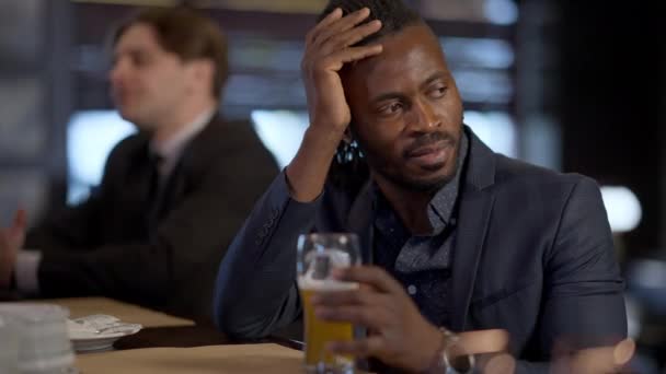 Portret Dotkniętego Afroamerykanina siedzącego przy barze ze szklanką piwa jako zamazanego białego faceta zamawiającego drinka w tle. Smutny zestresowany biznesmen w garniturze pijący w domu. — Wideo stockowe