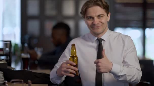 Retrato de vista frontal de un hombre caucásico seguro que muestra el pulgar hacia arriba sonriendo mirando a la cámara sosteniendo una botella de cerveza. Cliente alegre publicidad lager en el bar en el interior. — Vídeos de Stock