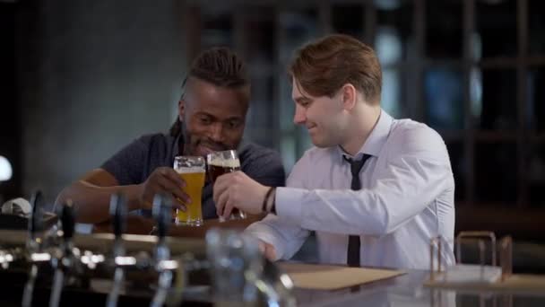Dos alegres y felices hombres relajados tintineando vasos de cerveza sonriendo tostando mirando a la cámara. Alegre amigos caucásicos y afroamericanos disfrutando de descansar en el bar el viernes por la noche en el interior. — Vídeo de stock