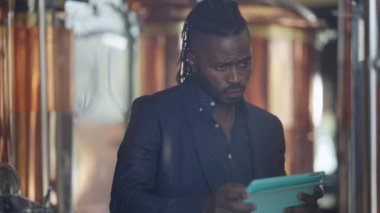 Ciddi bir Afro-Amerikan uzmanı büyük bira tankını tabletle kontrol ediyor. İçerideki bira fabrikasında alkol kalitesini denetleyen özgüvenli başarılı bir adamın portresi..