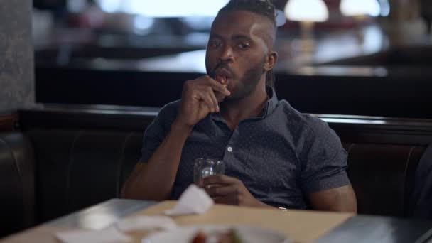 Красивый уверенный афроамериканец ест жареных куриных ножек, сидя с пивом в ресторане и разговаривая с неузнаваемым другом. Беззаботный парень отдыхает в помещении в пятницу вечером. — стоковое видео