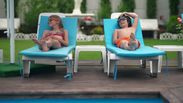 正面笑的白人男孩和女孩躺在木制游泳池甲板日光浴的日光浴床上。在户外度假的阳光明媚的日子里，欢乐的朋友们尽情享受暑假的全景. — 图库视频影像