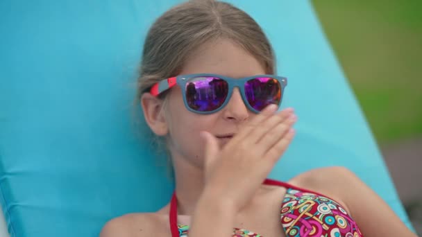 Großaufnahme Porträt eines glücklichen kaukasischen Mädchens mit Sonnenbrille, das in Zeitlupe Luftküsse sendet und lächelnd auf der Sonnenbank im Freien liegt. Charmante niedliche Kind posiert Sonnenbaden im Sommerresort an sonnigen Tag. — Stockvideo