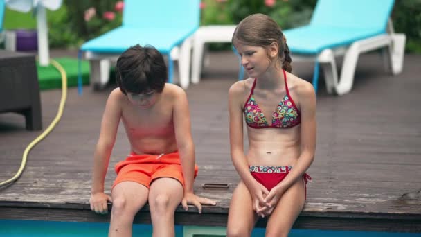 Okouzlující uvolněné děti sedící na dřevěné palubě u bazénu a flirtující. Přední pohled portrét bělocha chlapec a dívka mluví ve zpomaleném filmu těší letní volný čas venku na slunný den. — Stock video