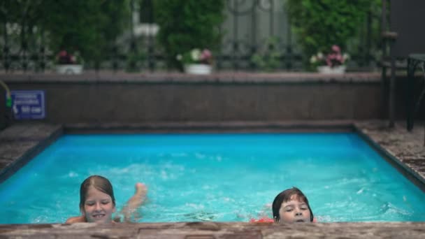 Alegre niño y niña nadando a la orilla de la piscina en cámara lenta mirando a la cámara y sonriendo. Retrato de alegre pareja caucásica en el fondo disfrutando de vacaciones de verano refrescantes en agua azul. — Vídeos de Stock