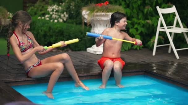 Wide shot of joyful couple of kids having fun playing with water gun sitting on poolside outdoors. Happy playful Caucasian boy and girl enjoying leisure on vacations on tourist resort on sunny day. — Stock Video