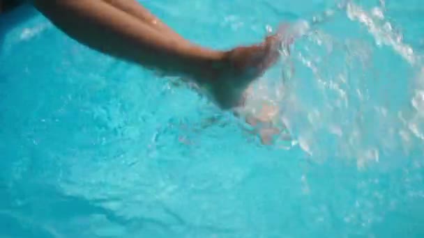 Közelkép vékony gyermek lábak fröccsenő azúrkék víz medencében lassított felvétel a napsütésben. Felismerhetetlen kaukázusi gyerek, aki jól érzi magát a medence mellett, üdülőhelyen. Gyermekkor és szabadidő. — Stock videók