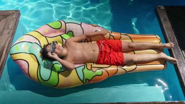 屋外プールでのスイミングマットレスの日光浴に横たわってサングラスでリラックスした小さな男の子のトップビュー。観光地で夏休みを楽しむ幸せな白人の子供の幅広いショット. — ストック動画