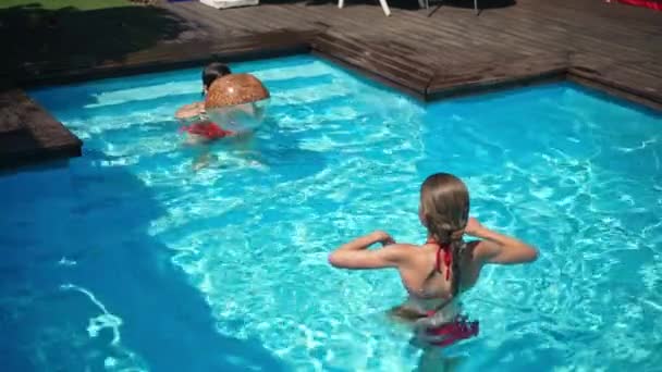 Niños jugando con pelota en la piscina en cámara lenta. Vista de ángulo alto de niño y niña caucásicos hiperactivos felices que se divierten disfrutando de las vacaciones de verano en el rayo de sol en agua azul. — Vídeo de stock