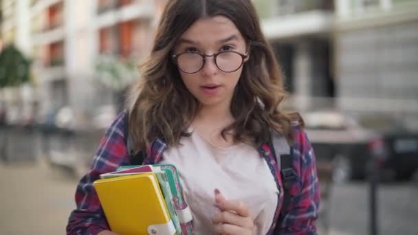 Vidám, magabiztos tinédzser lány, szemüveget igazít, mosolyog a kamerába. Egy magabiztos kocka kaukázusi egyetemista portréja, aki a város utcáján pózol. Bizalom és hírszerzés. — Stock videók