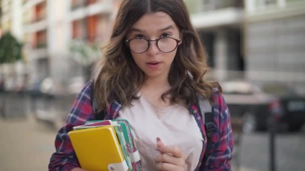 Alegre chica adolescente segura de ajustar las gafas sonriendo mirando a la cámara. Retrato de nerd confiado estudiante universitario caucásico posando en la calle de la ciudad. Concepto de confianza e inteligencia. — Vídeo de stock