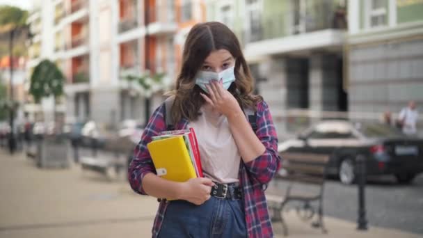 コロナウイルスのスマート魅力的な大学生の肖像画は本で街の通りにポーズマスクに直面しています。美しい白人の10代の少女が屋外でカメラを見て立っている。Covid-19の新ノーマル. — ストック動画