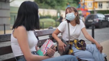 Covid-19 maskeli, sırt çantalı rahat bir üniversite öğrencisi elinde kitaplarla dışarıda oturmuş arkadaşlarıyla sohbet ediyor. Şehir caddesinde salgın hakkında konuşan Kafkas grup arkadaşları.