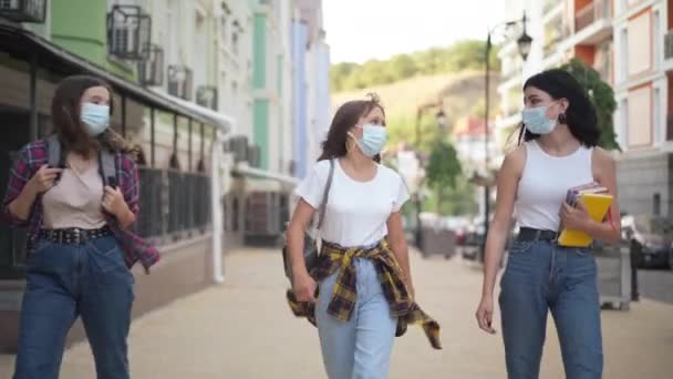 Retrato de tres estudiantes universitarios caucásicos en máscaras faciales Covid paseando por la calle de la ciudad con mochilas hablando. Chicas adolescentes seguras caminando al aire libre después de clases sobre la pandemia del coronavirus. — Vídeo de stock