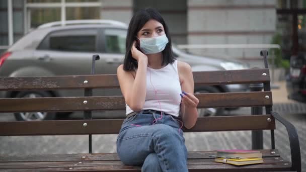 Pohled zepředu portrét smutné kavkazské dospívající dívky v Covid face masce poslouchající hudbu ve sluchátkách myšlení. Dospívající vysokoškolák těší koníček na ulici města na coronavirus pandemie ohniska — Stock video