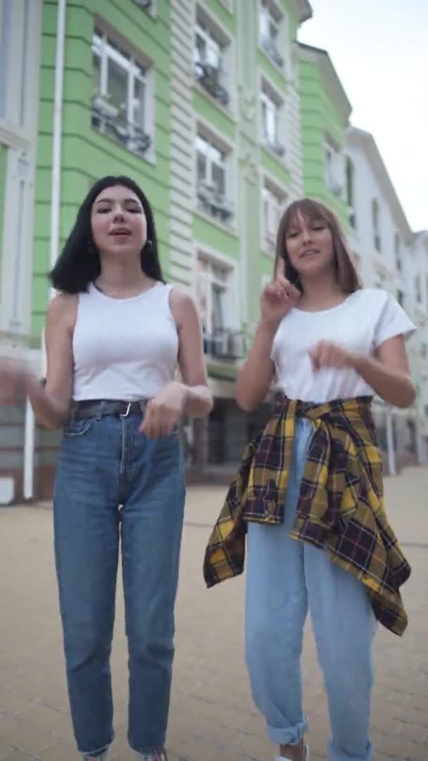 Vidám tinédzser lányok felveszik a táncot a közösségi médiának a City Streeten. Függőleges videó két pozitív vidám egyetemista táncáról, akik a szabadban nézik a kamerát. POV szelfi kamera. — Stock videók